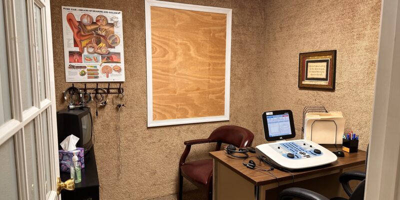 Small audiology test room with hearing test equipment, chairs, and ear anatomy poster on wall.