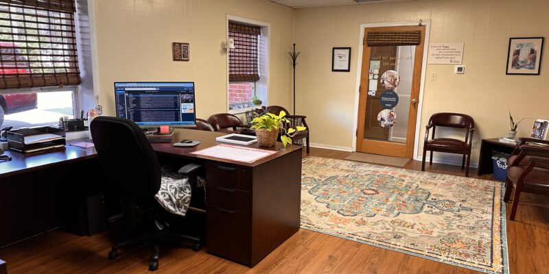 Office with wooden floors, desk with computer, chairs, plants, and a door with signage and window blinds.