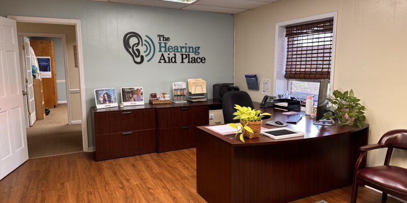 Reception area with desk, office chair, plants, and "The Hearing Aid Place" logo on the wall.