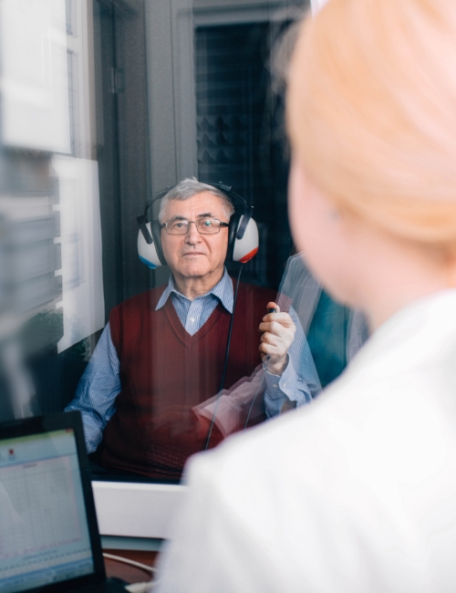 Older person wearing headphones during a hearing test with a healthcare professional visible in foreground.