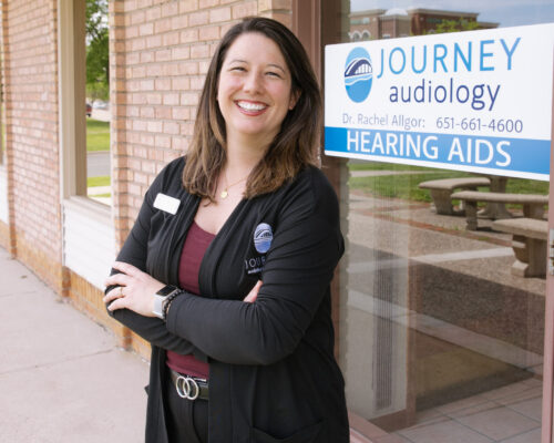 Smiling person standing outside Journey Audiology office next to a hearing aids sign on the door.