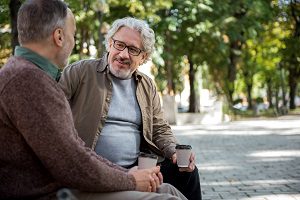 two men talking outside on a bench drinking coffee