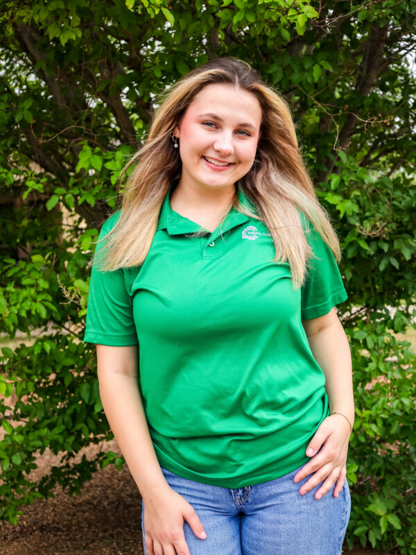Person smiling outdoors wearing a green polo shirt and blue jeans, standing in front of leafy bushes.