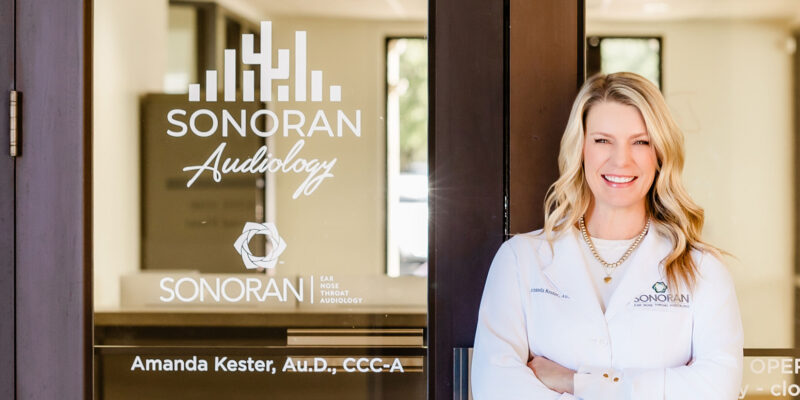 Amanda Kester, AuD standing outside of Sonoran Audiology hearing clinic with the sign visible on the door.