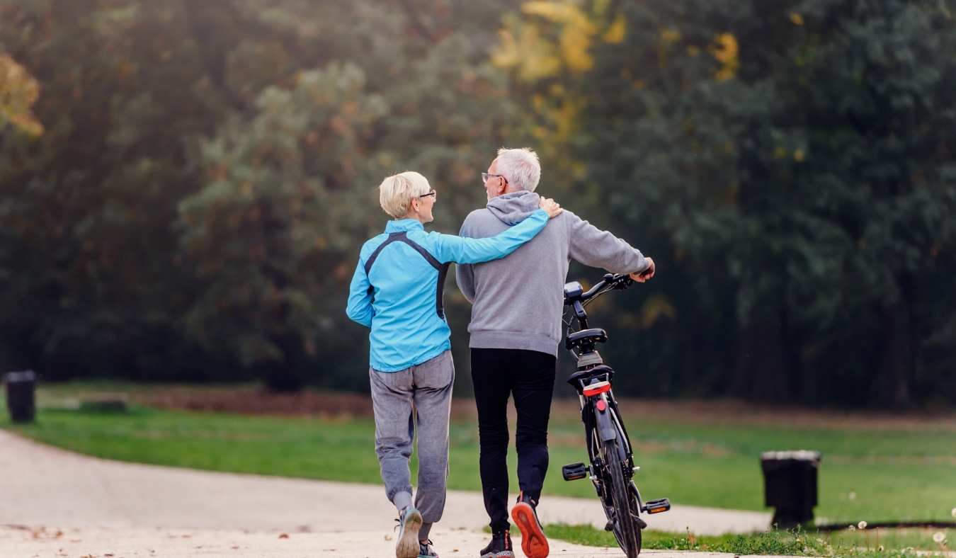 Cheerful active senior couple with bicycle walking through park together