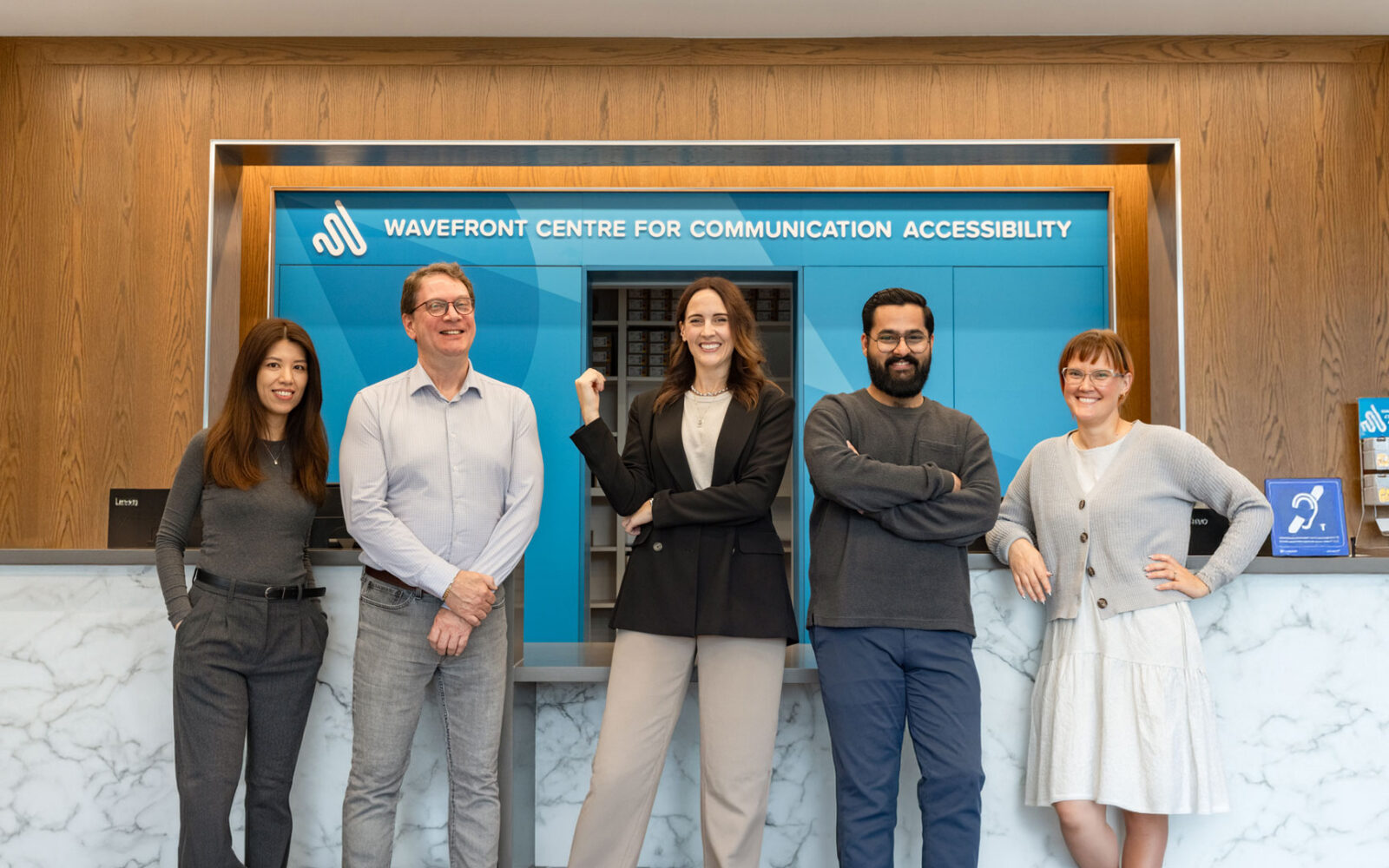 Five members of the Wavefront Hearing team smile at the reception desk beneath a sign reading “Wavefront Centre for Communication Accessibility.