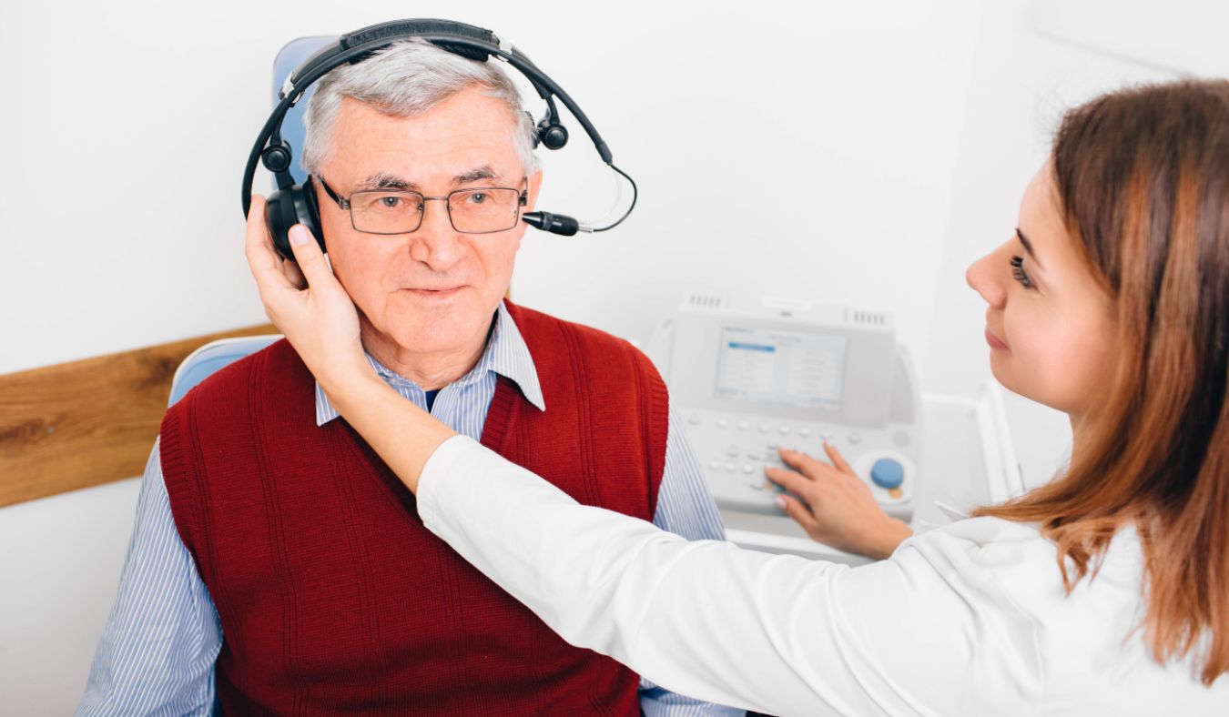 Senior man undergoing a hearing test with an audiologist adjusting headphones in a clinic setting.