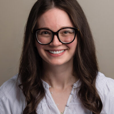 Person with long brown hair, black glasses, and white embroidered blouse smiling against a plain background.