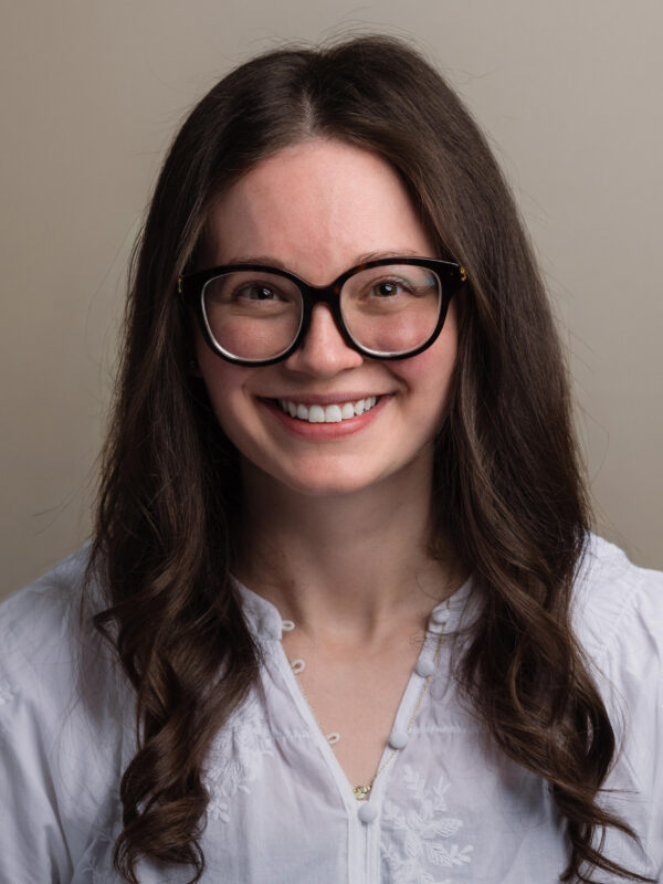 Person with long brown hair, black glasses, and white embroidered blouse smiling against a plain background.
