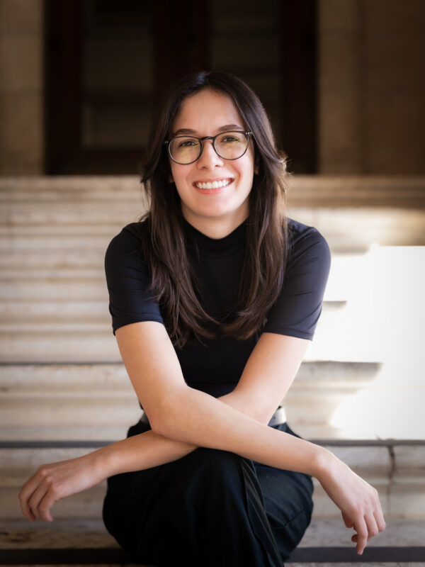 Person with long dark hair and glasses smiling, seated on stone steps with arms crossed.