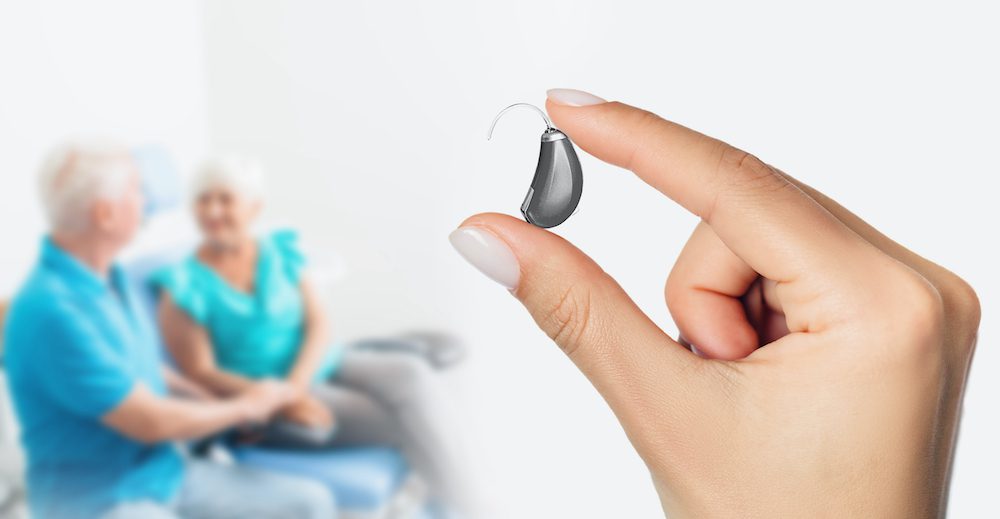 hand holding a silver hearing aid with two patients in the background