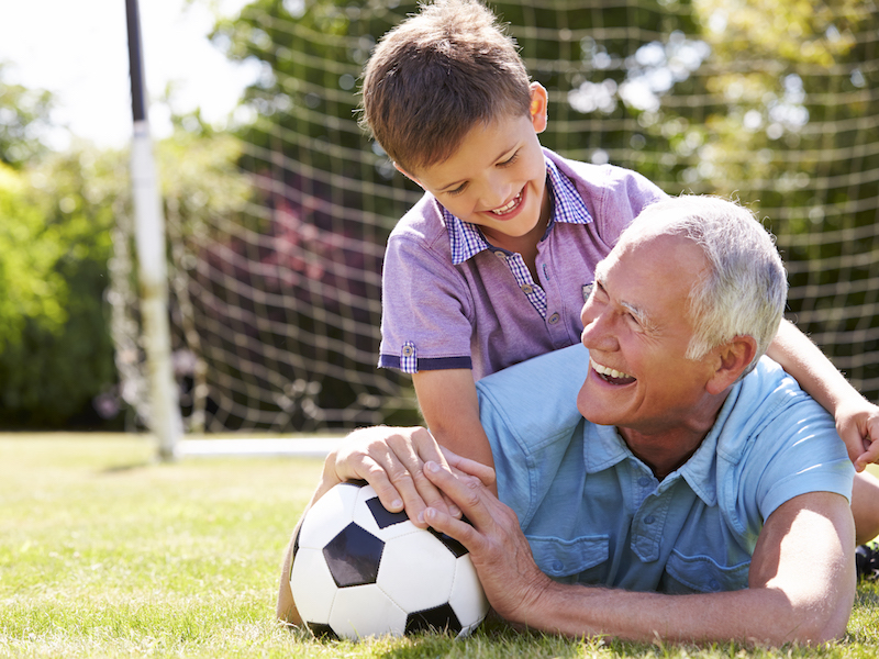 A man with hearing aids playing soccer with his grandson in a youthful way.