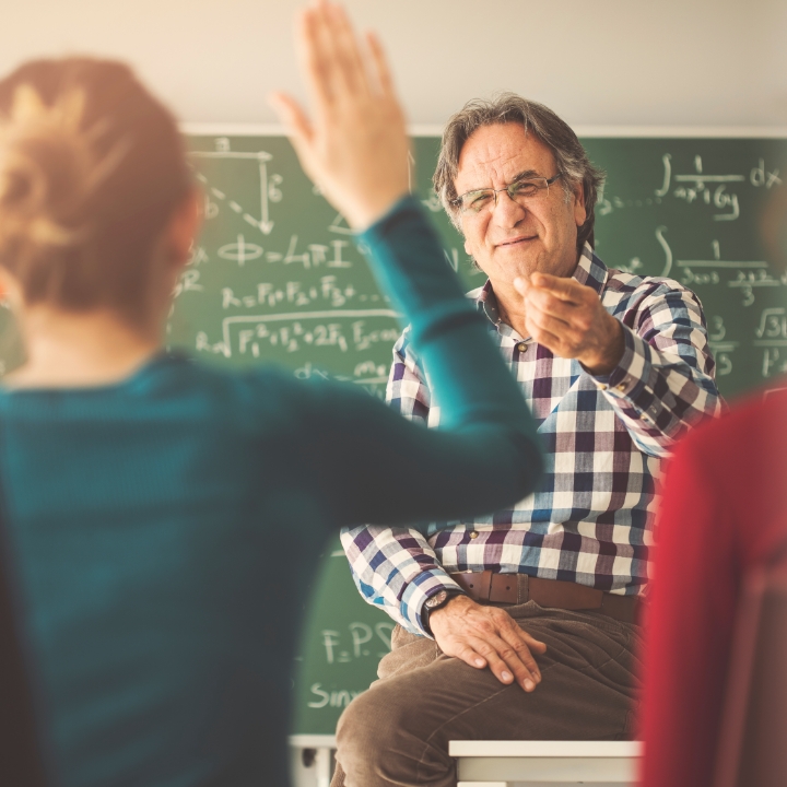 Teacher engaging with student raising hand in classroom with math equations on chalkboard.