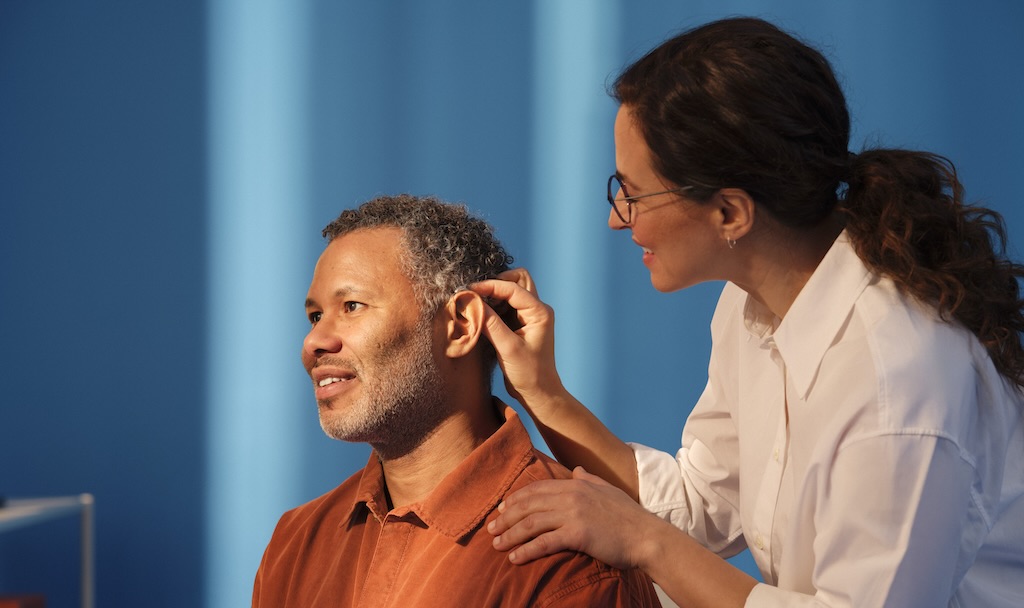 beltone boost ultra Person fitting a hearing aid into another person's ear in a clinical setting with blue background.