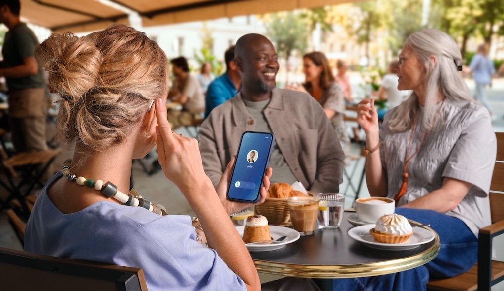 Person holding phone showing incoming call while sitting at outdoor café table with two others.