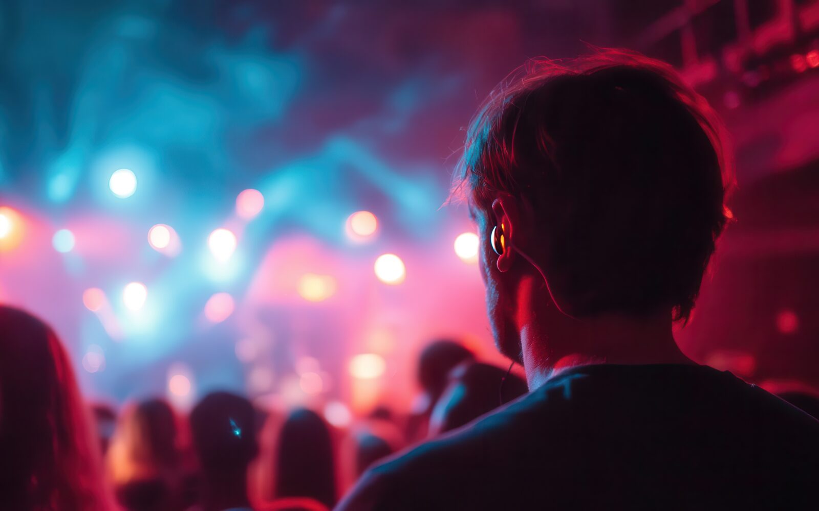 A man wearing earplugs watches a vibrant concert, surrounded by colorful stage lights and a lively crowd