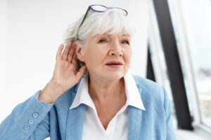 Come again please? Attractive mature woman wearing blue jacket and spectacles on her head eavesdropping Stylish retired female in elegant clothes holding hand at her ear, suffering from hearing loss