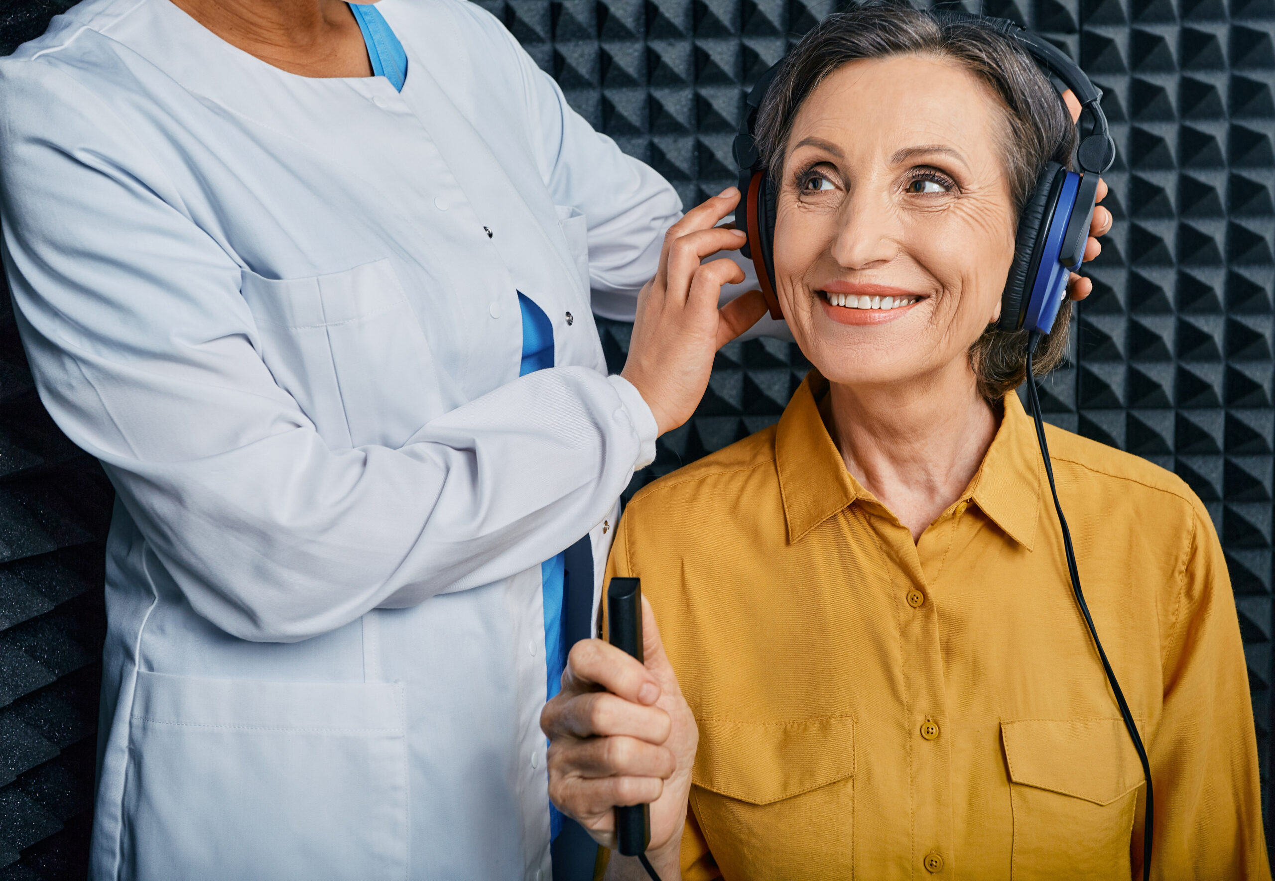 Portrait senior woman with white toothy smile while hearing check up with ENT doctor at soundproof audiometric booth using audiometry headphones and audiometer