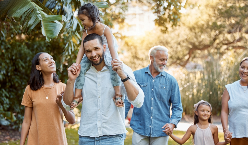 Mixed-age family walking outdoors on a sunny day, with a child on a man's shoulders.