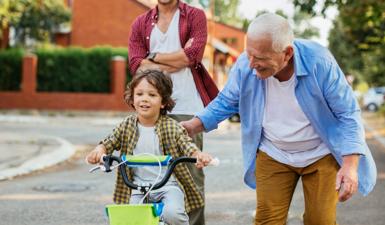 grandfather teaching grandson how to ride a bike with father in background
