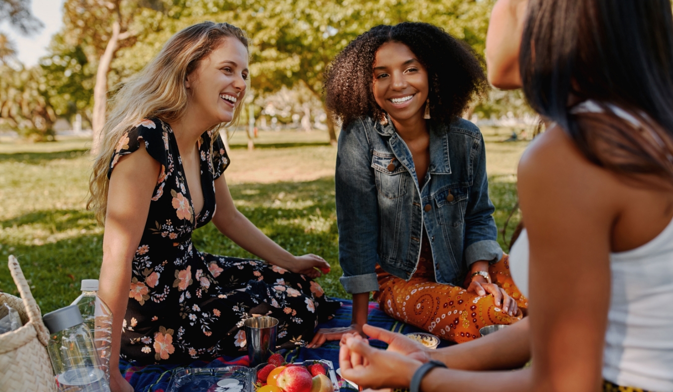 Group of smiling multiracial female best friends sitting together on blanket with fruits