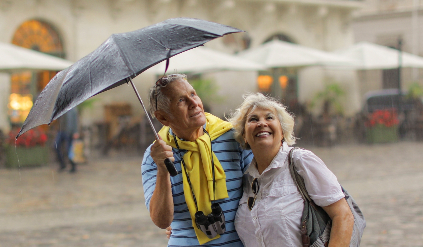 Happy senior two male and female tourists stand downtown and enjoy the rainy weather