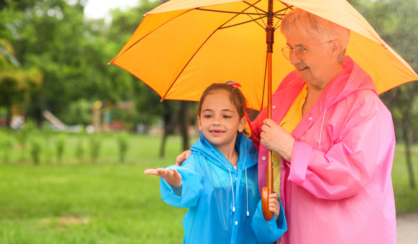 Little girl and her grandma in raincoats with umbrella outdoors