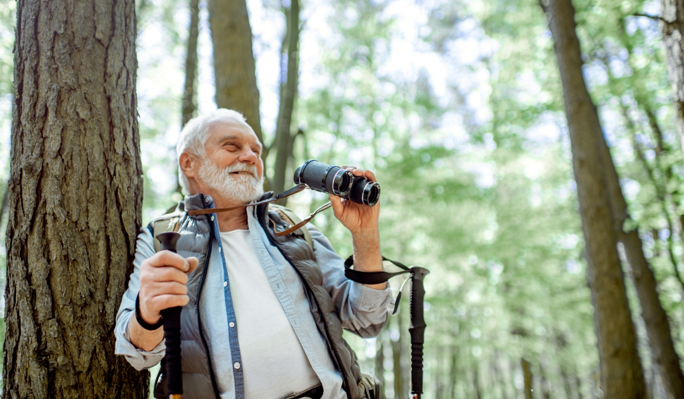 senior man with binoculars and backpack resting near the tree while traveling in the forest