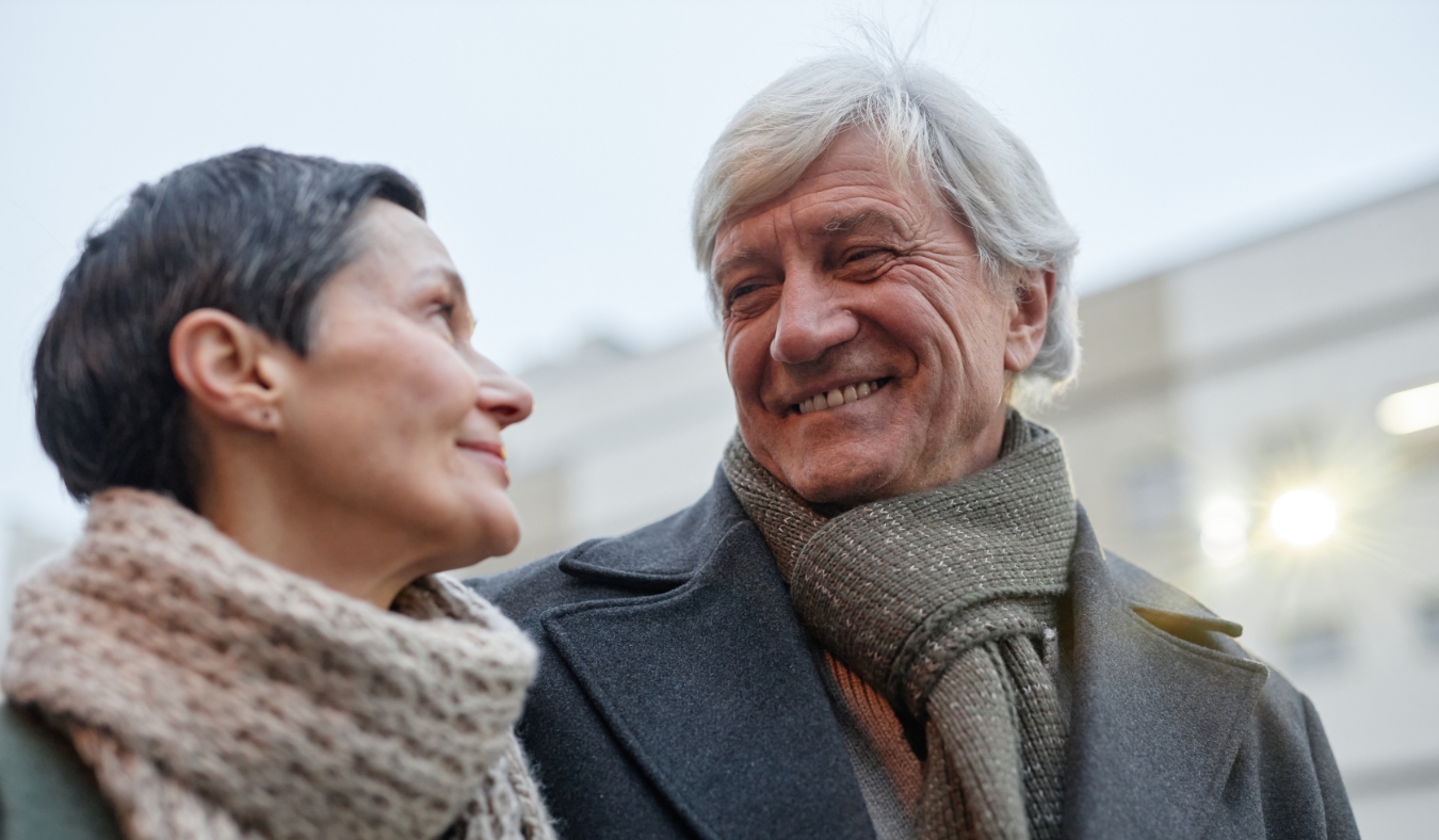 elderly couple smiling warmly at each other while wearing scarves and winter coats